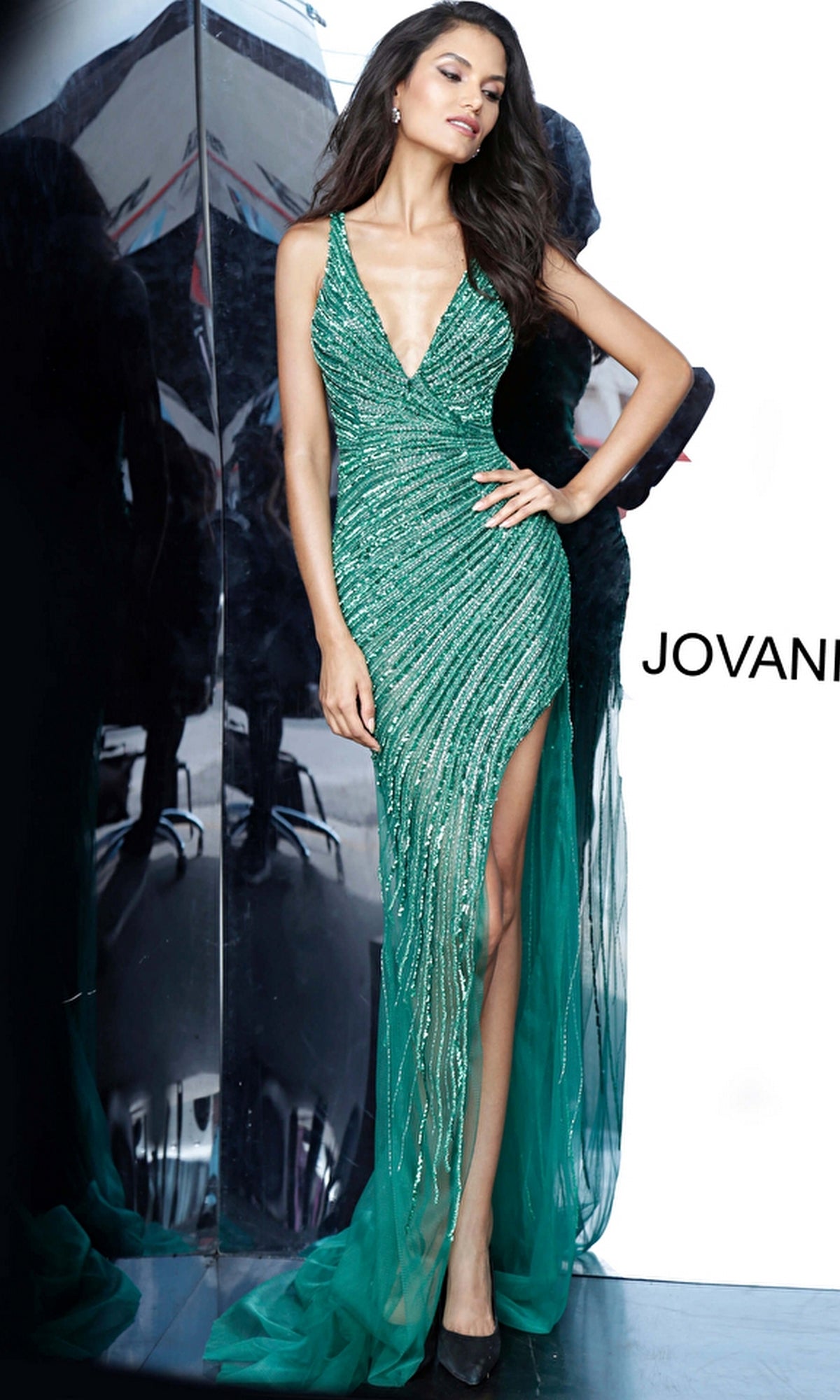 A woman in the Jovani 63405 Beaded-Mesh Sexy Long Prom Dress, featuring a sleeveless green sequined design with a deep V-neck and high slit, stands before a mirrored wall.